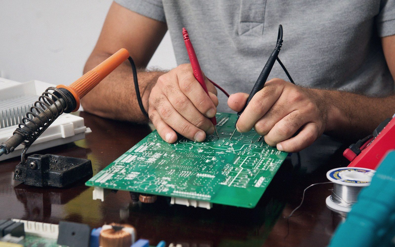 Hands using multimeter probes to test a green circuit board on a workbench